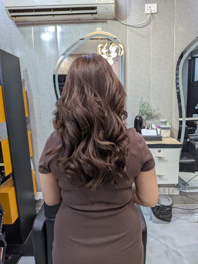 The back of a client's head showing long, loosely curled and highlighted hair, styled at a mirror station with a chandelier reflected in the glass.